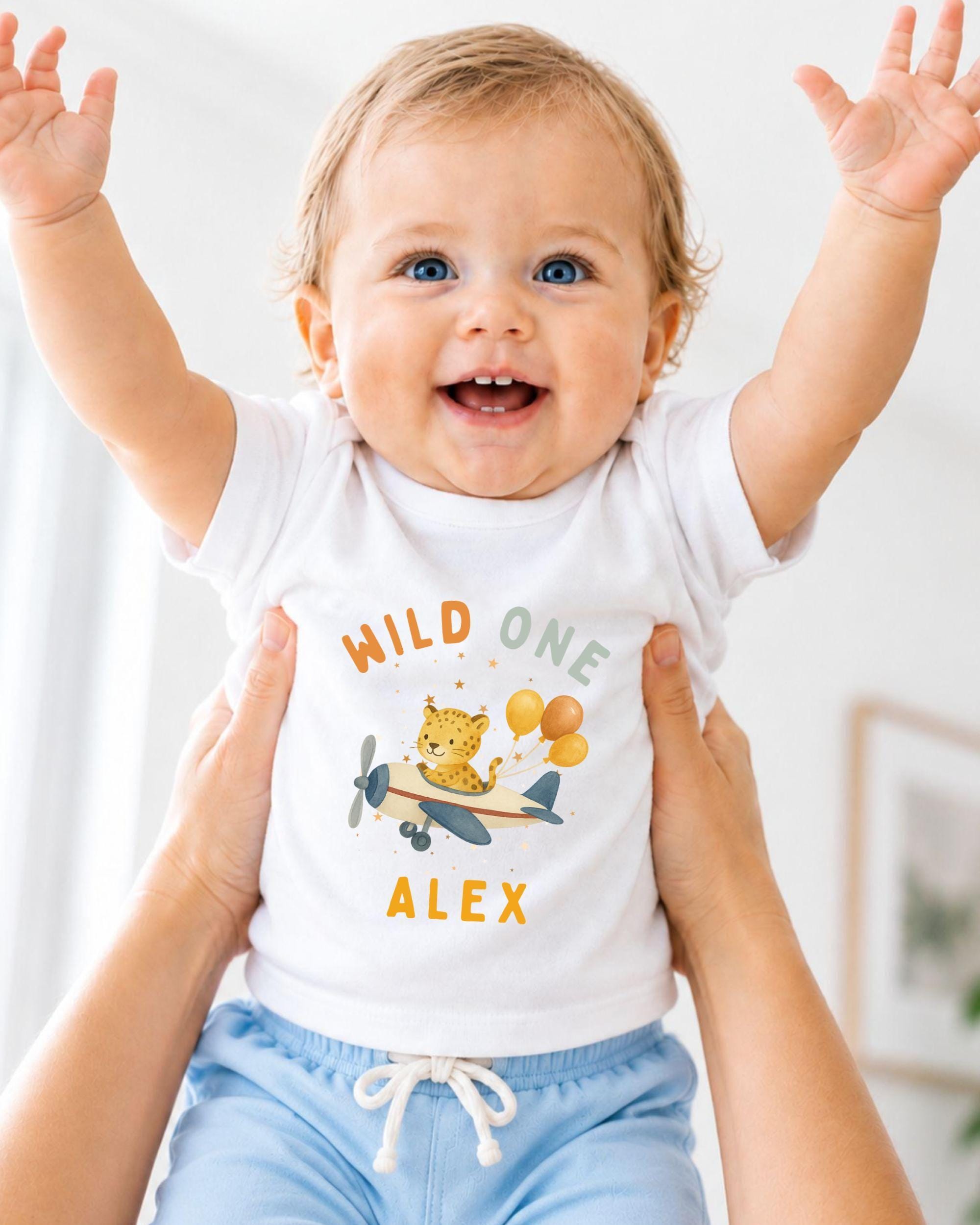 A smiling baby wearing a white t-shirt with a cartoon airplane and the name "Alex" printed on it, being held up by someone's arms.