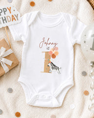 A white onesie with a zebra and balloons is displayed on a white blanket, surrounded by birthday decorations and a "Happy Birthday" sign.