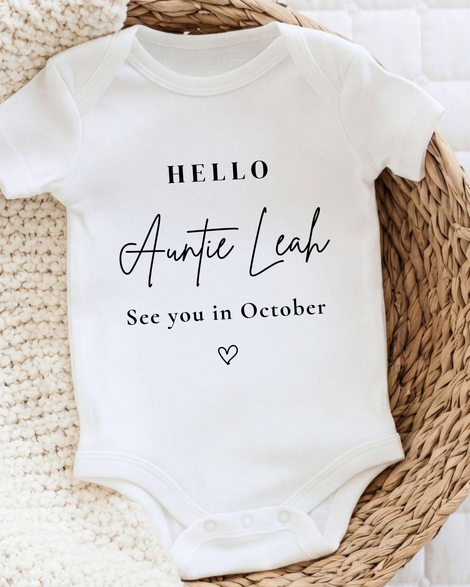A white baby onesie with the text "Hello Auntie Leah See you in October" printed on it, placed on a woven basket.