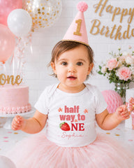 A young girl wearing a birthday hat and a white t-shirt with the text "half way to one" and "Sophia" is posing in front of a birthday cake and balloons.