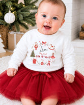 A smiling baby girl wearing a white sweater with a Christmas-themed design and a red tulle skirt, sitting on a white carpet in front of a Christmas tree.
