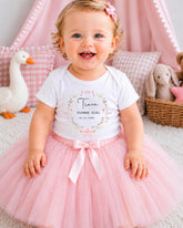 A smiling baby girl wearing a white t-shirt with a floral design and a pink tulle skirt, sitting on a pink carpet surrounded by stuffed animals.