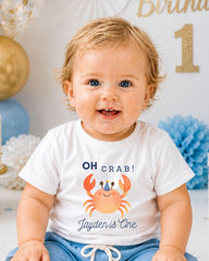 A smiling baby boy wearing a white t-shirt with a crab design and the text "OH Crab! Jayden is One" is sitting in front of a birthday party backdrop.