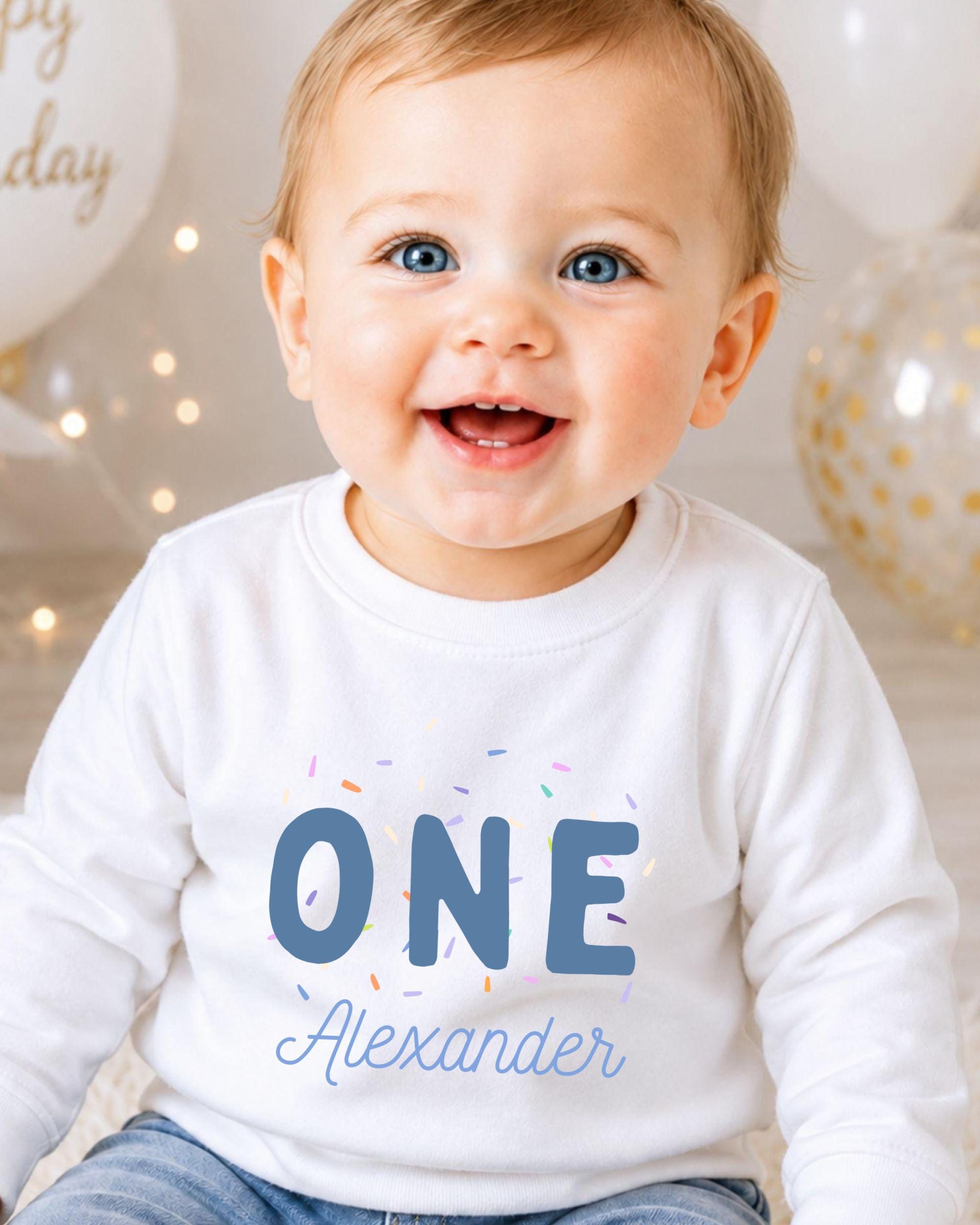 A smiling baby boy wearing a white shirt with the text "ONE Alexander" printed on it.