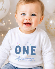 A smiling baby boy wearing a white shirt with the text "ONE Alexander" printed on it.