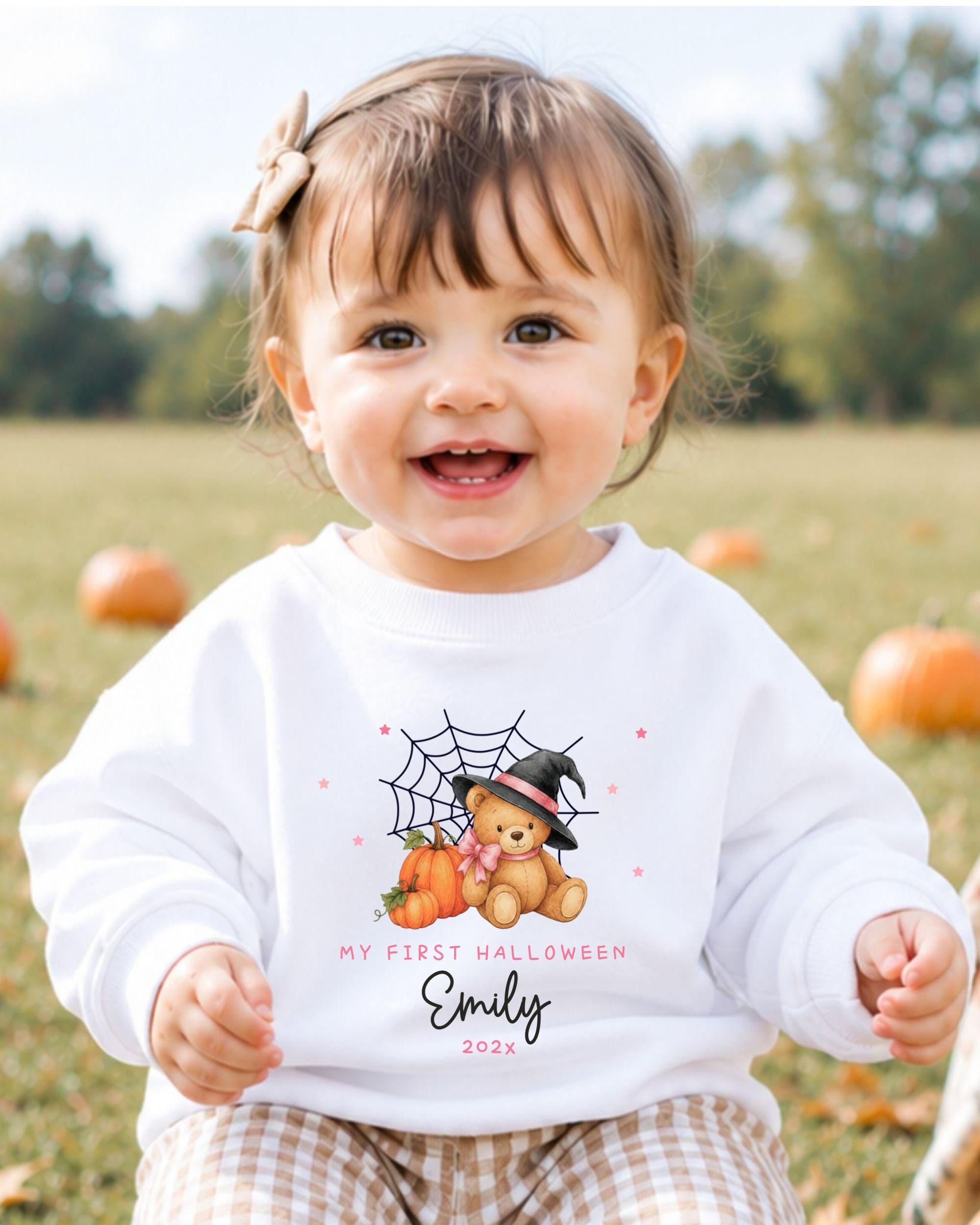 A smiling baby girl wearing a white sweater with a Halloween-themed design and text.