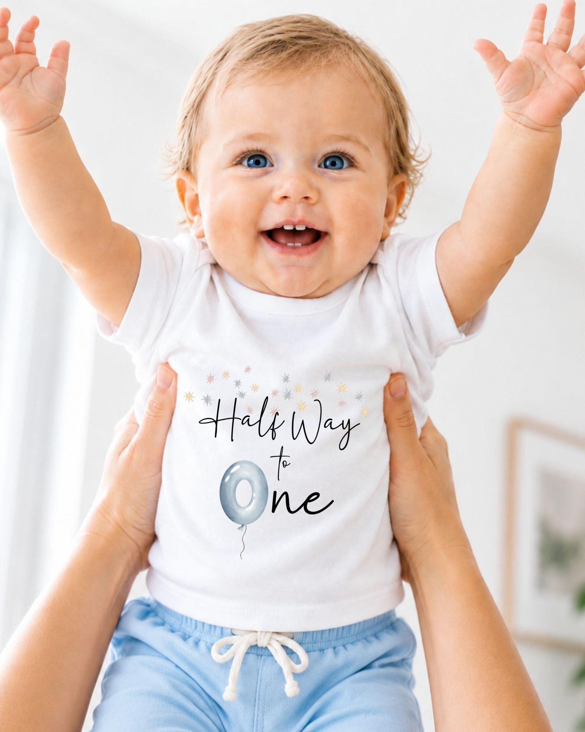 A smiling baby wearing a white t-shirt with the text "Half Way to One" and a balloon graphic, being held up by someone's arms.