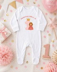 A white onesie with a princess and the number 1 printed on it, surrounded by pink and gold decorations, including a pink star, confetti, and a pink ribbon.