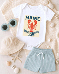 A white onesie with a lobster design, a beige bucket hat, and light blue shorts are arranged on a beige surface, along with sunglasses and seashells.