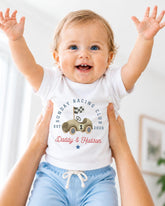 A smiling baby wearing a white t-shirt with a graphic of a race car and the text "Sunday Racing Club EST. 1926" is being held up by two people.