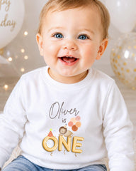 A smiling baby boy wearing a white shirt with a birthday message and a cartoon character.