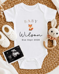 A baby announcement t-shirt with the name "Baby Wilson" and the date "Due Sept 2028" printed on it, along with a crocheted rabbit toy and a baby bootie.