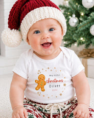 A smiling baby wearing a Santa hat and a white t-shirt with a gingerbread man graphic and text that says "My first Christmas Oliver 20x".