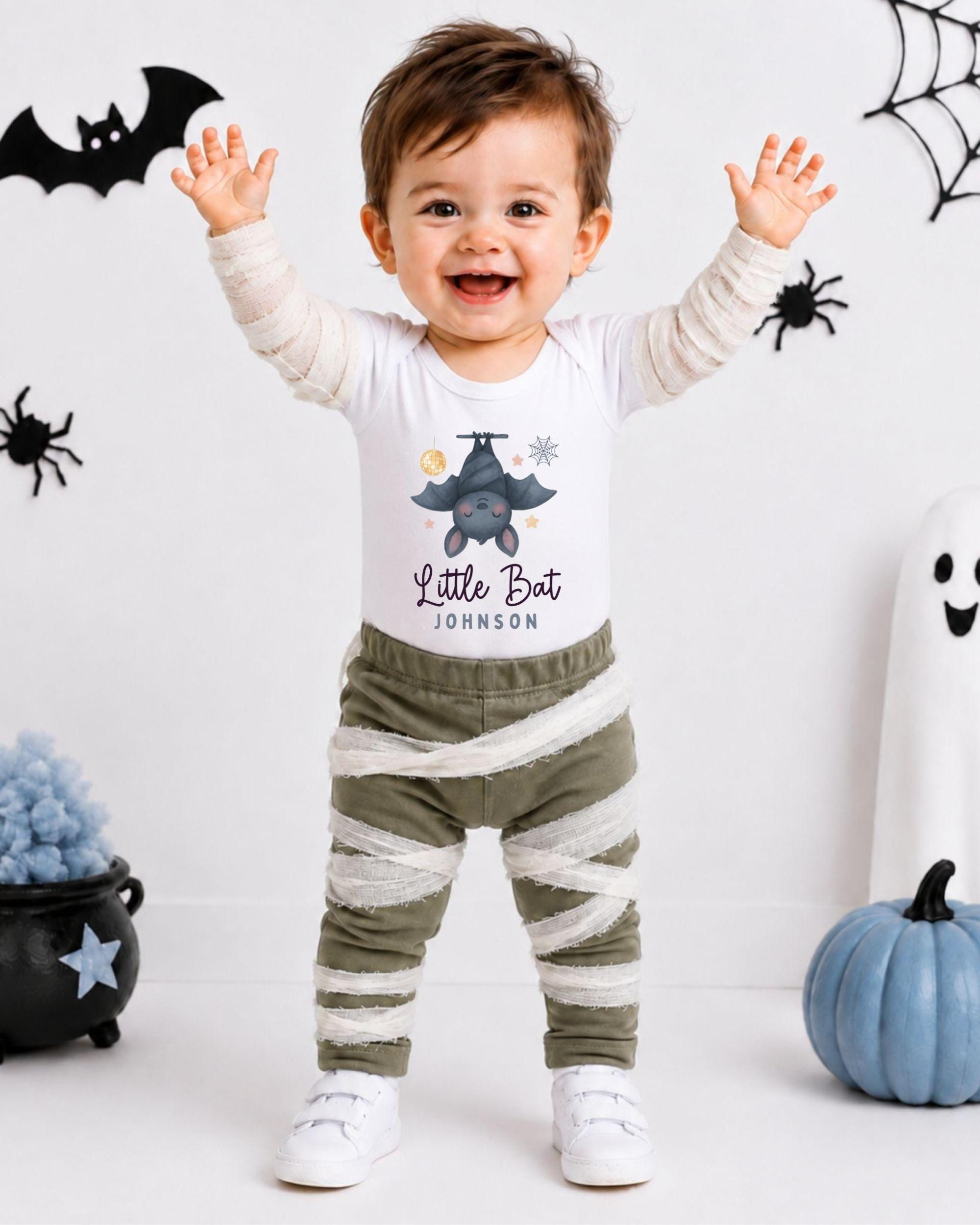 A smiling baby boy wearing a white t-shirt with a bat design and striped pants, standing in front of Halloween decorations.