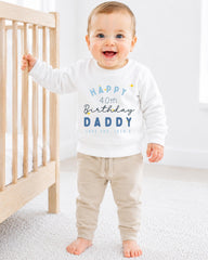 A smiling baby boy wearing a white sweatshirt with text and a birthday message, standing next to a wooden crib.
