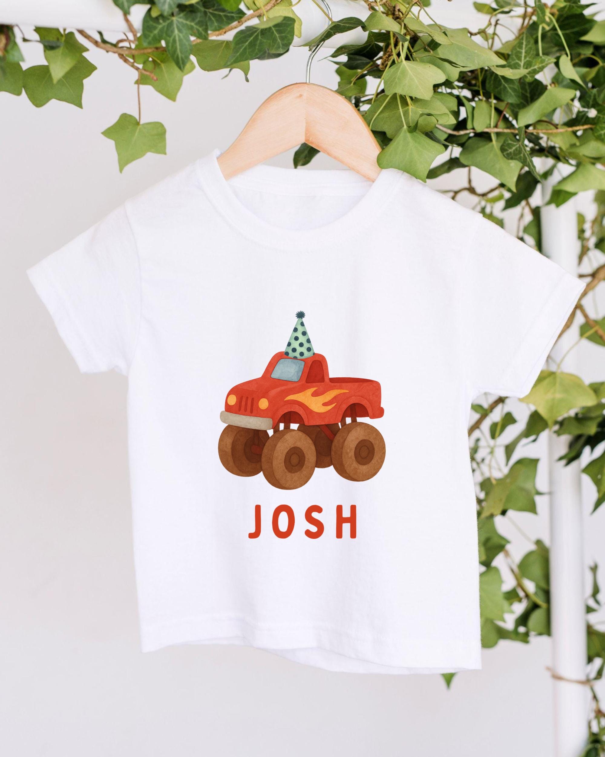 A white t-shirt with a red and orange monster truck graphic printed on it, hanging on a wooden hanger.