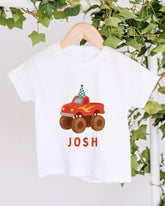 A white t-shirt with a red and orange monster truck graphic printed on it, hanging on a wooden hanger.