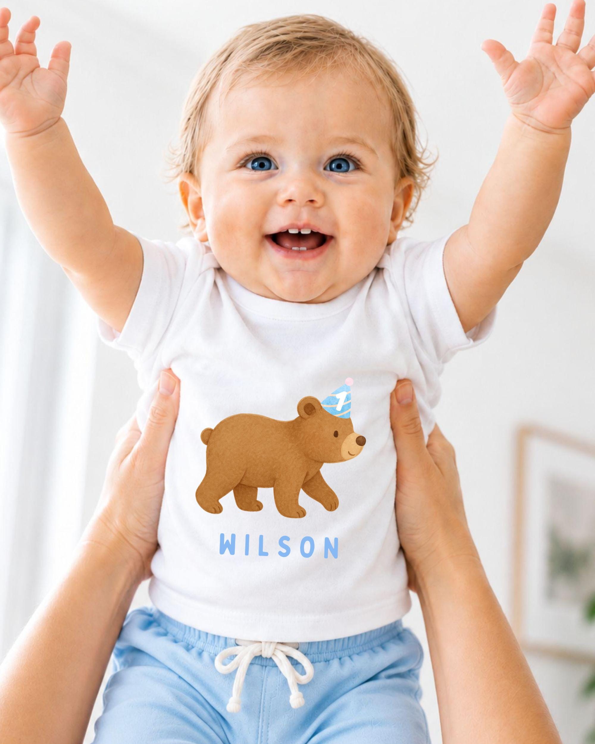 A smiling baby wearing a white t-shirt with a bear graphic and the name "WILSON" printed on it, being held up by someone's arms.