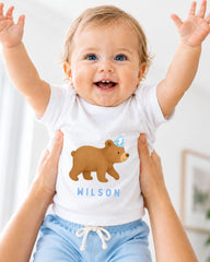 A smiling baby wearing a white t-shirt with a bear graphic and the name "WILSON" printed on it, being held up by someone's arms.