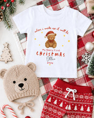 A white t-shirt with a teddy bear design, a knit hat, and red shorts, all arranged on a plaid blanket with Christmas decorations.