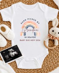 A white baby onesie with a rainbow graphic and text, along with a crocheted rabbit toy and a baby's firstborn photo, all arranged on a woven surface.