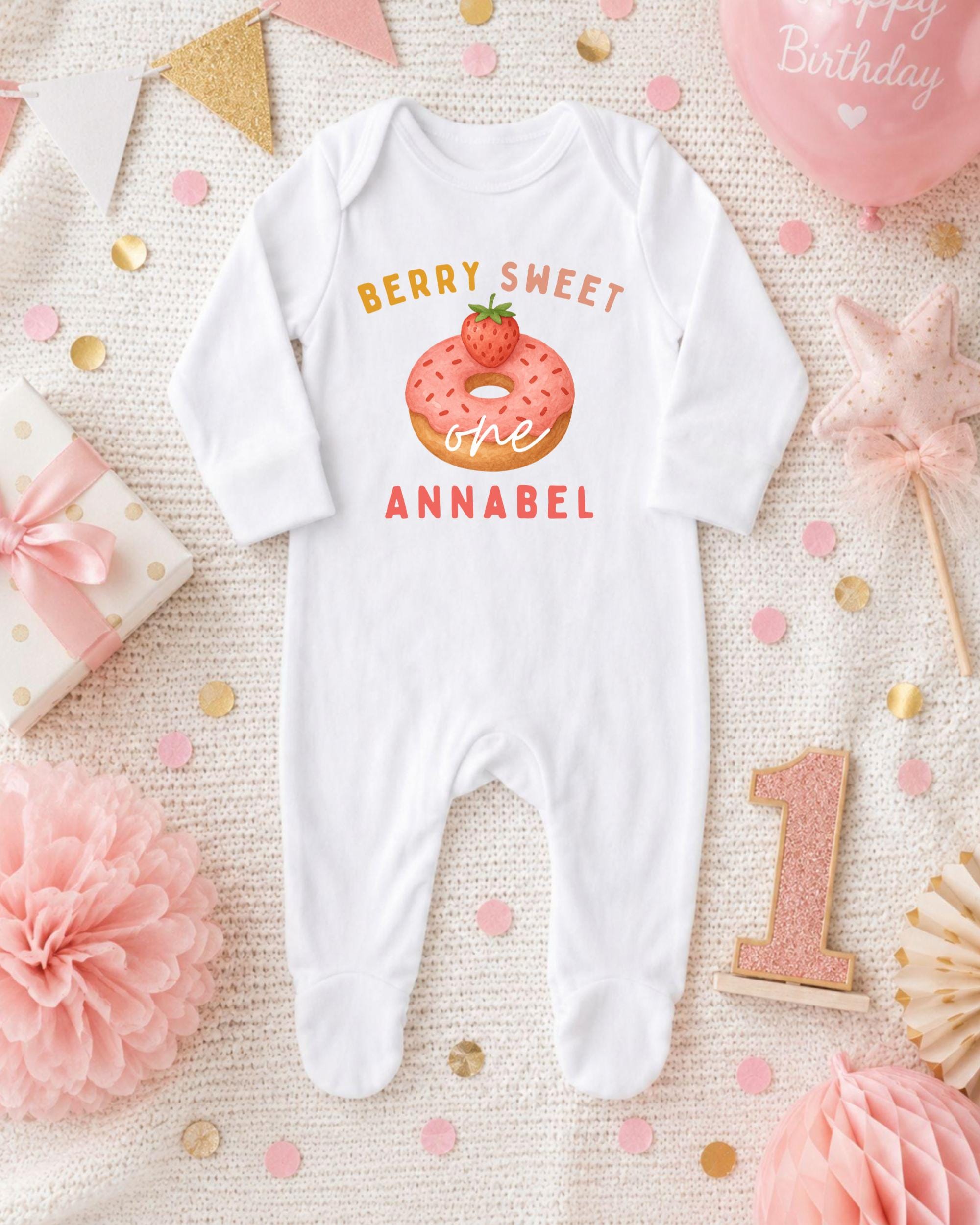 A white onesie with a pink donut and the text "Berry Sweet One Annabel" printed on it, surrounded by pink and gold confetti, a pink star, and a pink and gold birthday balloon.