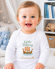 A smiling baby boy wearing a white onesie with a Viking ship design and the name "Vincent" printed on it, sitting in a room with a teddy bear and bookshelf in the background.