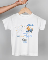 A white t-shirt with a graphic design featuring a cartoon character flying in a plane and the text "UP, UP & AWAY! FIRST TIME FLYER Oscar 2026".