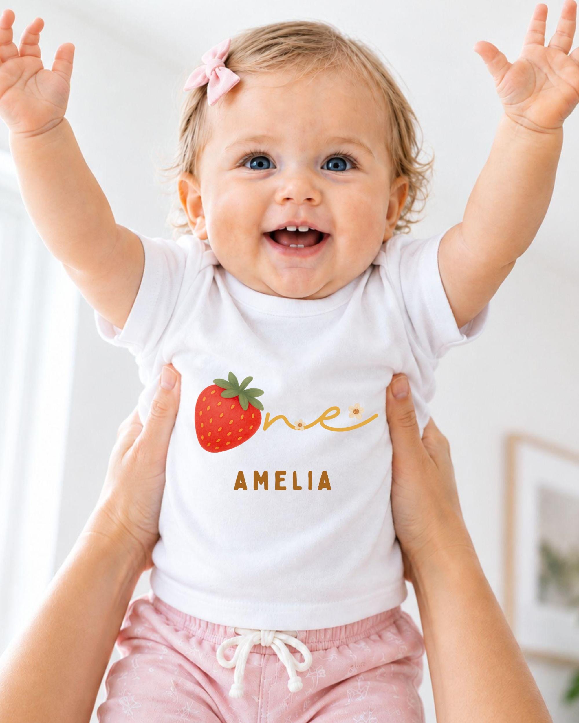 A smiling baby girl wearing a white t-shirt with a strawberry design and the name "Amelia" printed on it, being held up by someone's arms.