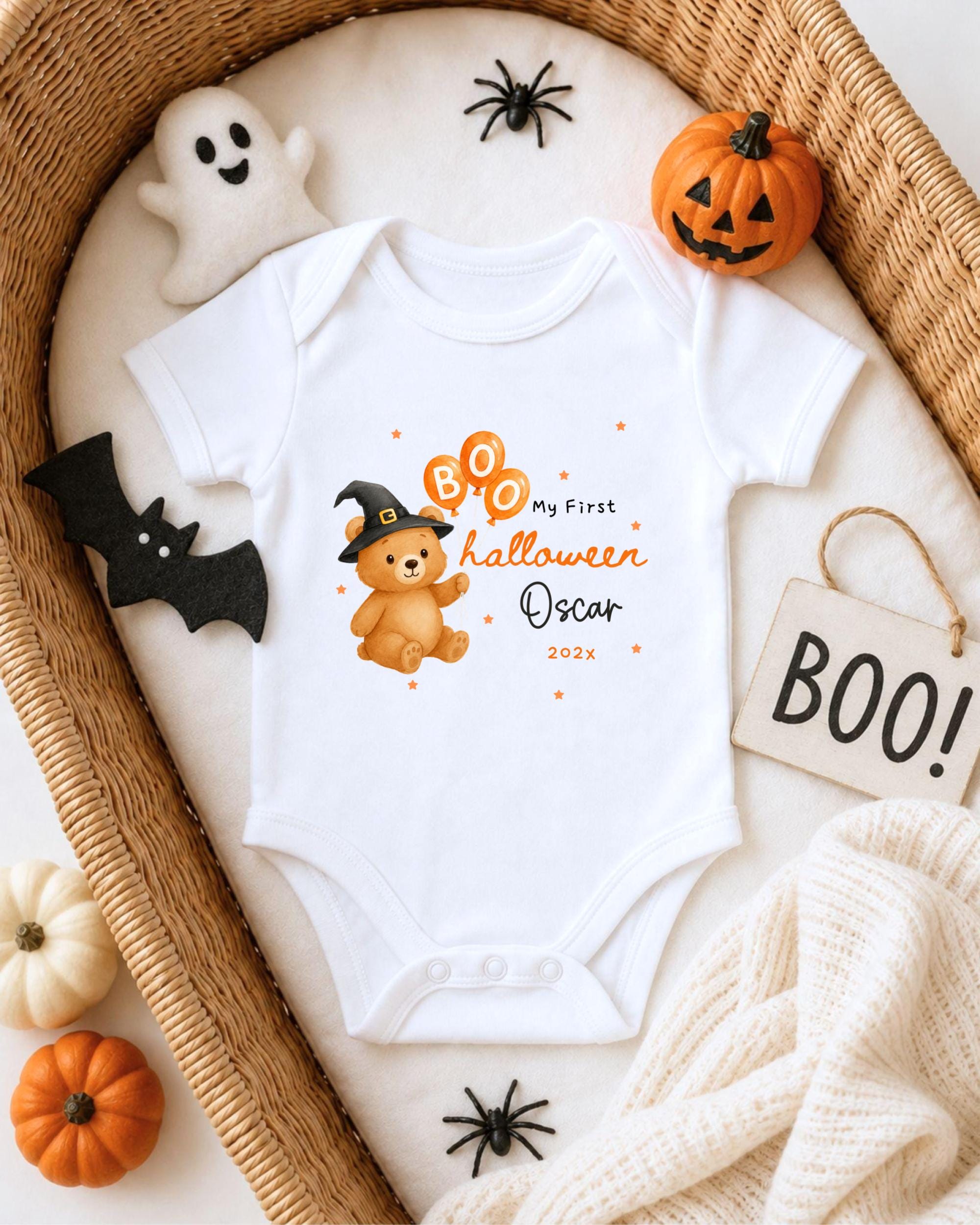 A white onesie with a teddy bear and a witch hat, along with Halloween decorations such as a pumpkin and a ghost, are arranged on a bed.
