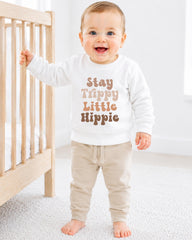 A smiling baby boy wearing a white sweatshirt with the text "Stay Trippy Little Hippie" printed on it, standing next to a wooden crib.