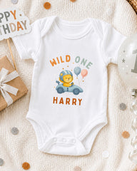 A white onesie with a cartoon lion and the text "WILD ONE HARRY" printed on it, along with a birthday sign and balloons, suggesting a celebration for a baby's first birthday.