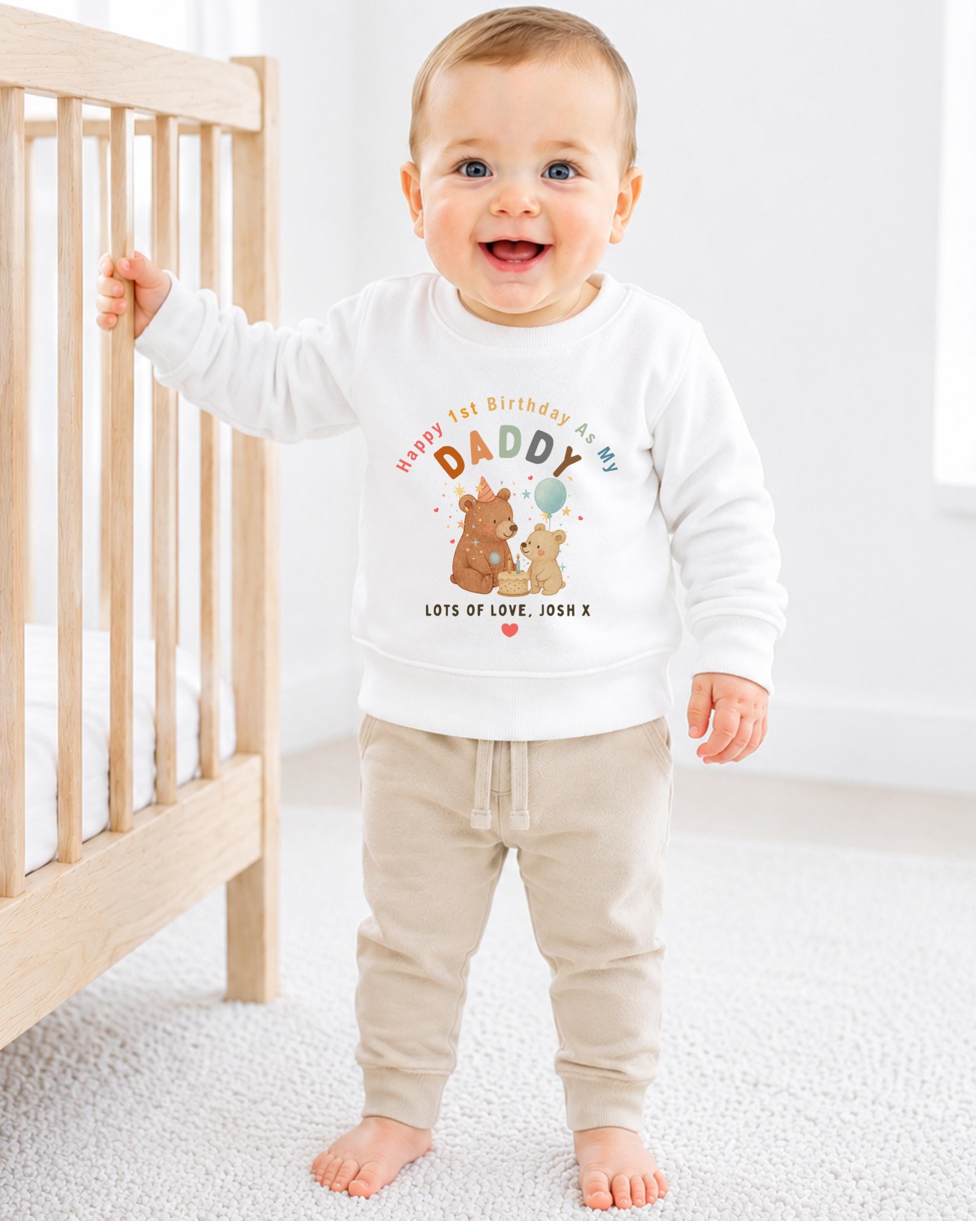 A smiling baby boy wearing a white sweatshirt with a cartoon image of a bear and the text "Happy 1st Birthday As My Daddy Lots of Love, Josh X" is standing in front of a wooden crib.