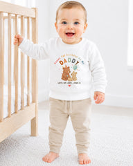 A smiling baby boy wearing a white sweatshirt with a cartoon image of a bear and the text "Happy 1st Birthday As My Daddy Lots of Love, Josh X" is standing in front of a wooden crib.