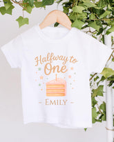 A white t-shirt with a graphic design featuring a slice of cake and stars, along with the text "Halfway to One - Emily" printed on it.