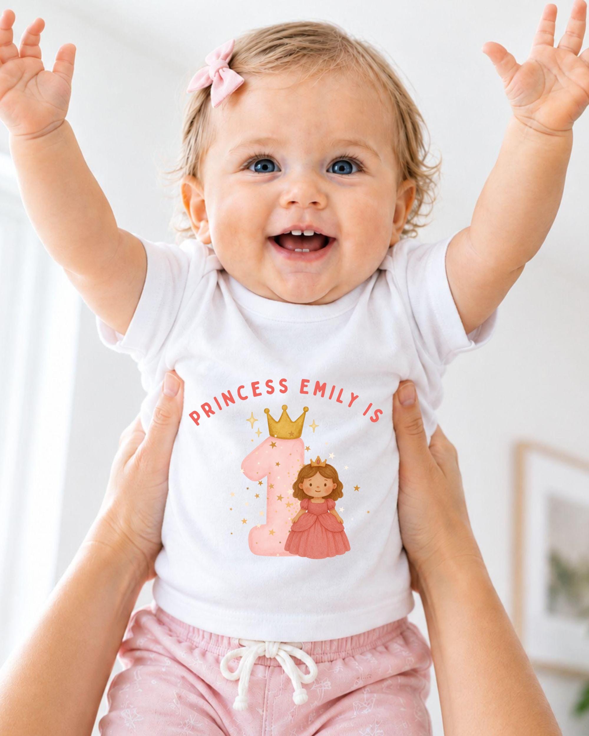 A smiling baby girl wearing a white t-shirt with a princess and number 1 design, being held up by someone's arms.