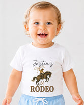 A smiling baby boy wearing a white t-shirt with a cowboy graphic and text that says " Justin's 1st Rodeo".