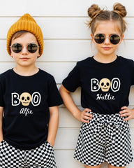 Two children wearing black t-shirts with the text "BOO" and "Alfie" printed on them, along with skull designs, stand against a white wall.