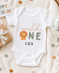 A white onesie with a lion design and the text "wild one leo" printed on it, along with a "Happy Birthday Leo" sign and a birthday balloon.