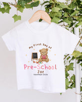 A white t-shirt with a teddy bear and a chalkboard, along with the text "My First Day of Pre-School Zoe September 2020".