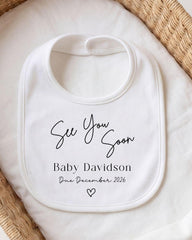 A white baby bib with the text "See You Soon Baby Davidson Due December 2026" printed on it.