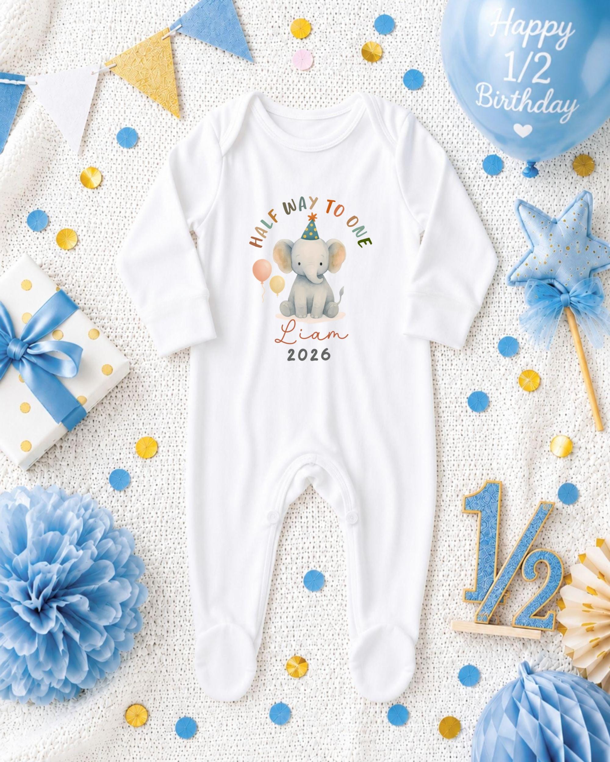 A white onesie with a cartoon elephant and the text "Half way to one" is displayed among various baby-themed items, including a blue balloon, confetti, and a star.