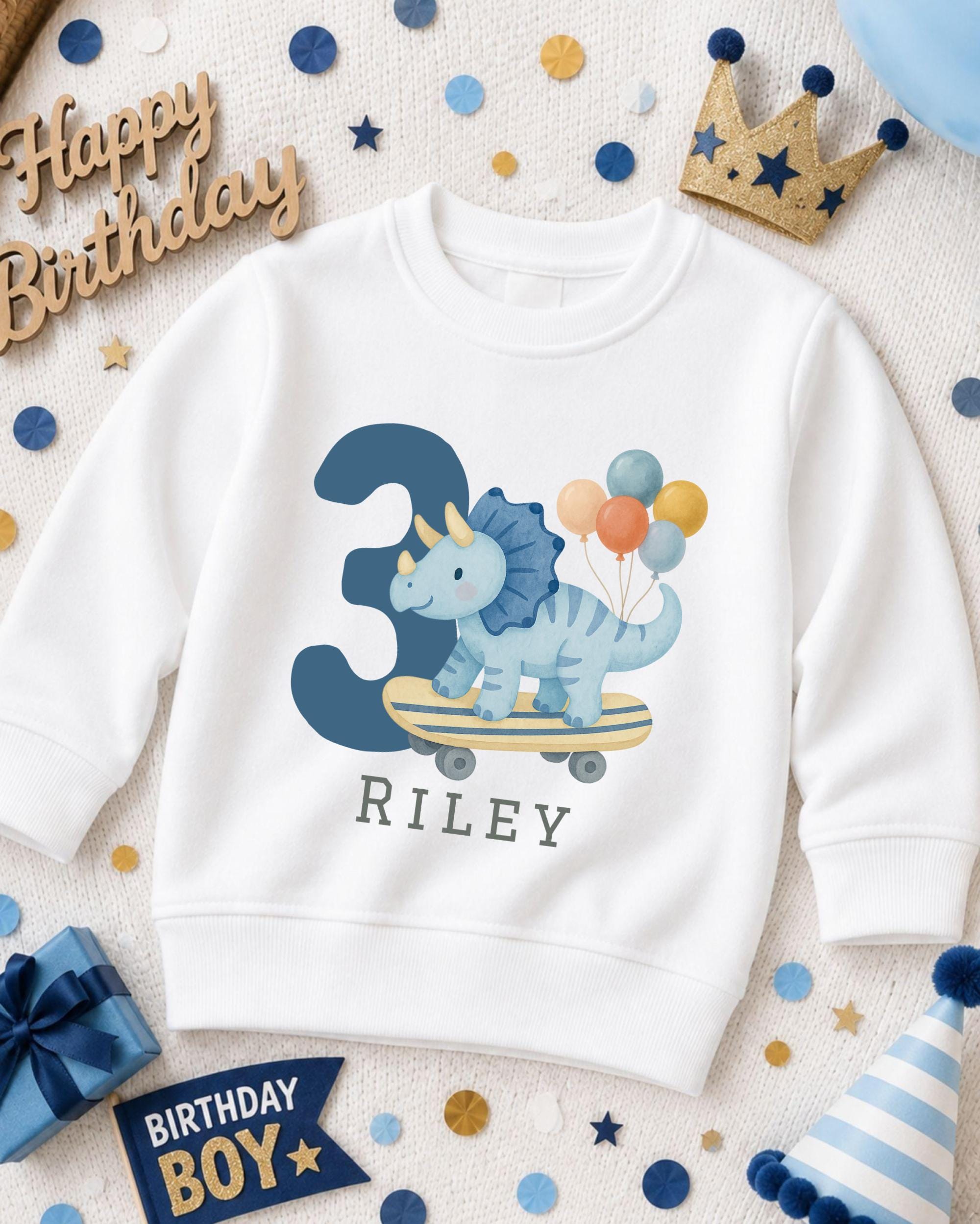 A white sweatshirt with a dinosaur riding a skateboard and the number 3 printed on it, surrounded by birthday decorations and a birthday hat.