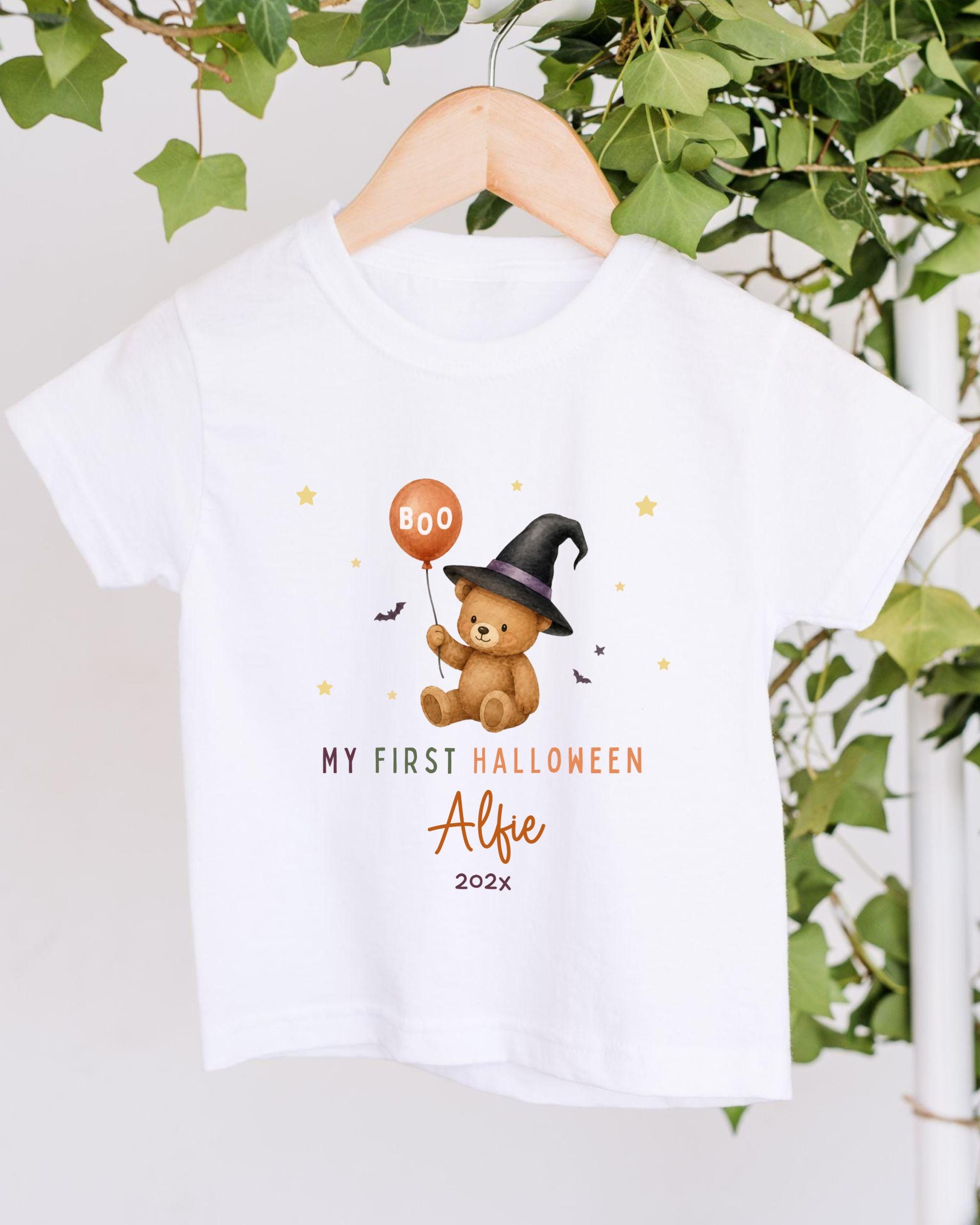 A white t-shirt with a graphic design featuring a teddy bear wearing a witch's hat and holding a "boo" balloon, along with the text "My first Halloween Albie 2020x".