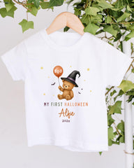 A white t-shirt with a graphic design featuring a teddy bear wearing a witch's hat and holding a "boo" balloon, along with the text "My first Halloween Albie 2020x".