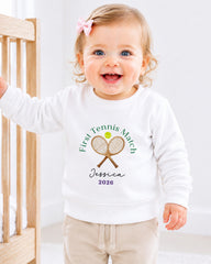 A smiling baby girl wearing a white sweatshirt with a tennis racket and ball design, standing next to a wooden crib.