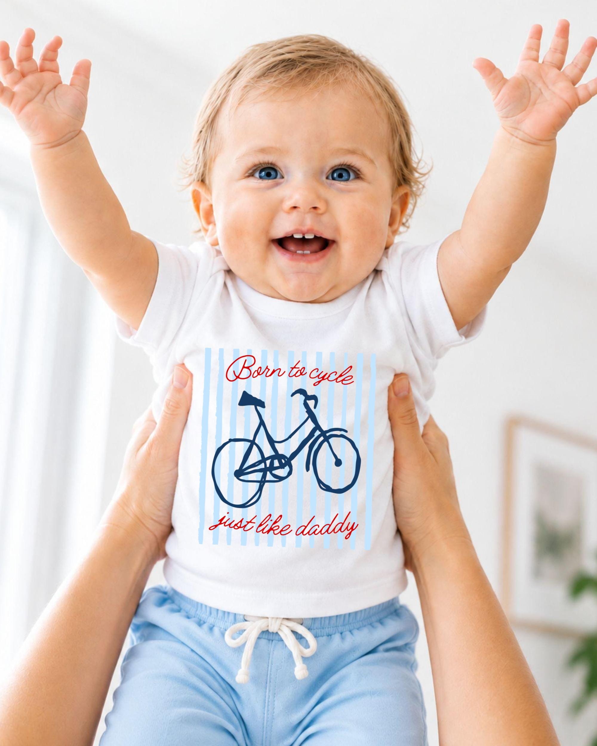 A smiling baby wearing a white shirt with a bicycle graphic and the text "Born to cycle just like daddy" is being held up by someone's arms.