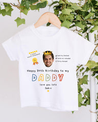 A white t-shirt with a personalized message and a crown image, hanging on a wooden hanger.