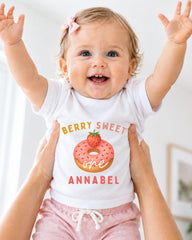 A smiling baby girl wearing a white t-shirt with a strawberry donut graphic and the text "Berry Sweet one Annabel" is being held up by someone's arms.