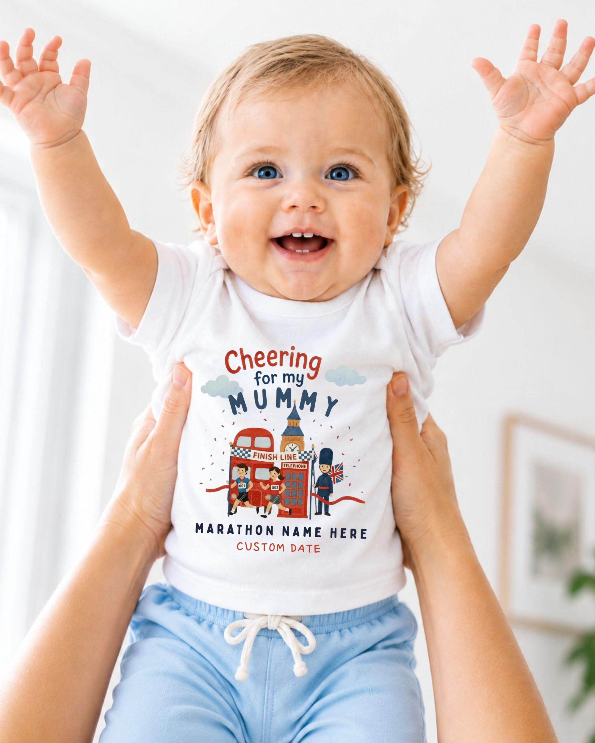 A smiling baby wearing a white t-shirt with a graphic design and text, being held up by someone's arms.